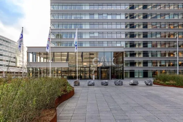 Vooraanzicht van het CAK-gebouw aan de Prinses Beatrixlaan met glazen entree, vlaggen en bestrate ingang met grote stenen.